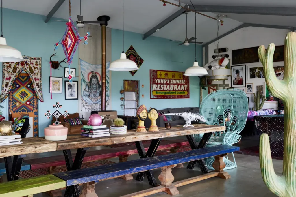 A vibrant eclectic maximalist room featuring a long rustic wood dining table with mismatched painted benches, colorful global textiles, bold wall art, a turquoise peacock chair, and playful decor accents in a high-ceiling open space.