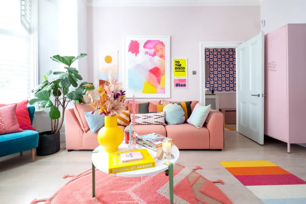 A colorful eclectic maximalist living room with a blush pink sofa, vibrant pillows, abstract art, a yellow vase with dried flowers, and a blue accent chair. The space includes color-blocked decor, playful wall art, and a round white coffee table with books and candles.