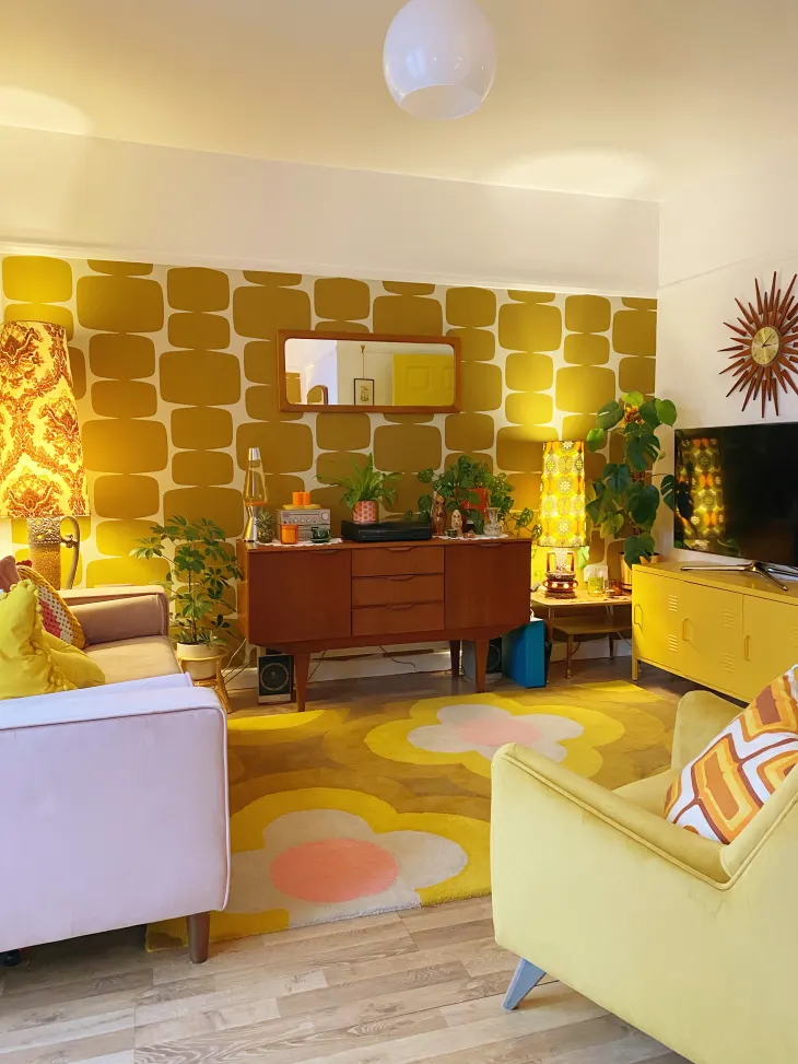 Eclectic maximalism living room with bold retro wallpaper, vibrant yellow and pink furniture, mid-century decor, floral area rug, and layered lighting elements.